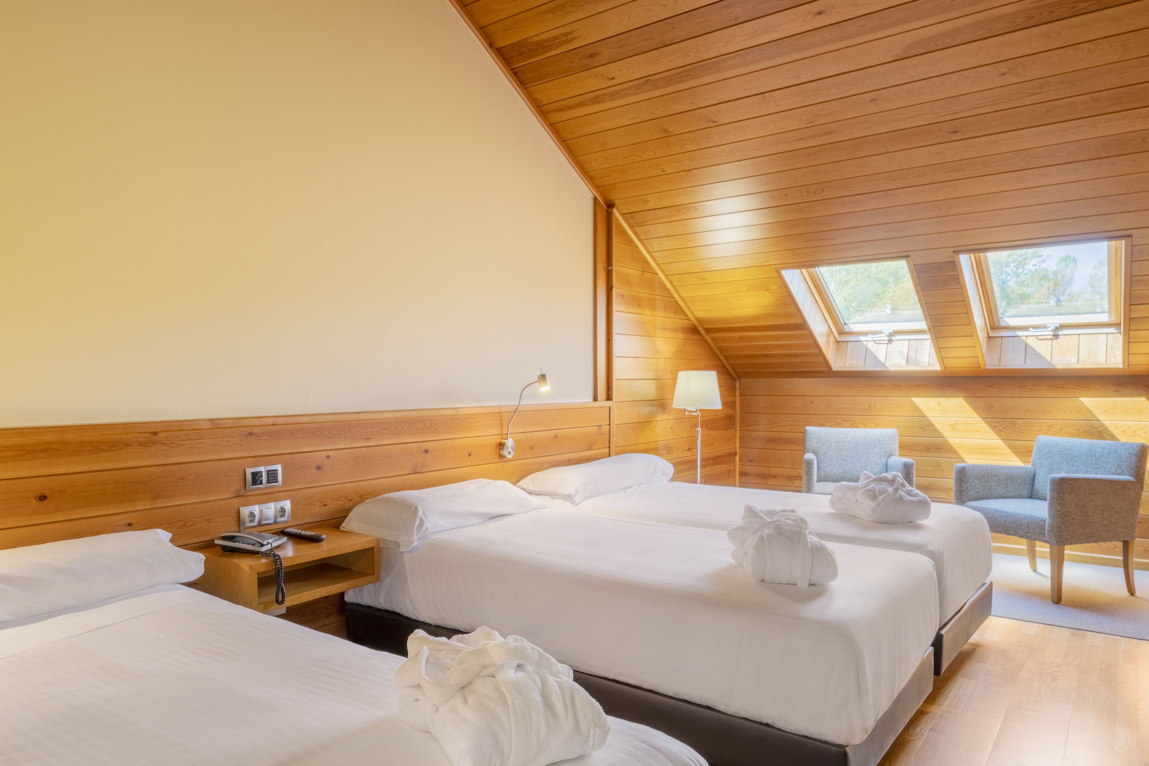Habitación de hotel con dos camas, techo de madera, tragaluces, una mesita de noche con lámpara y una silla.