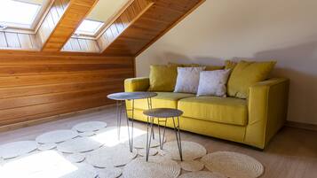 A room with a sloped ceiling, wooden walls, a yellow sofa with cushions, two small round tables, and a patterned rug.