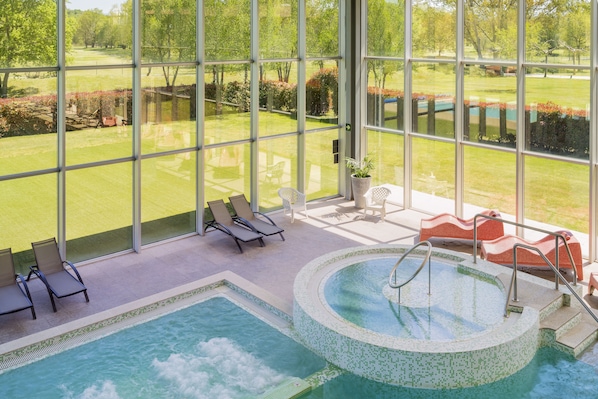 A pool area with lounge chairs, a hot tub, and a view of a golf course through large windows.