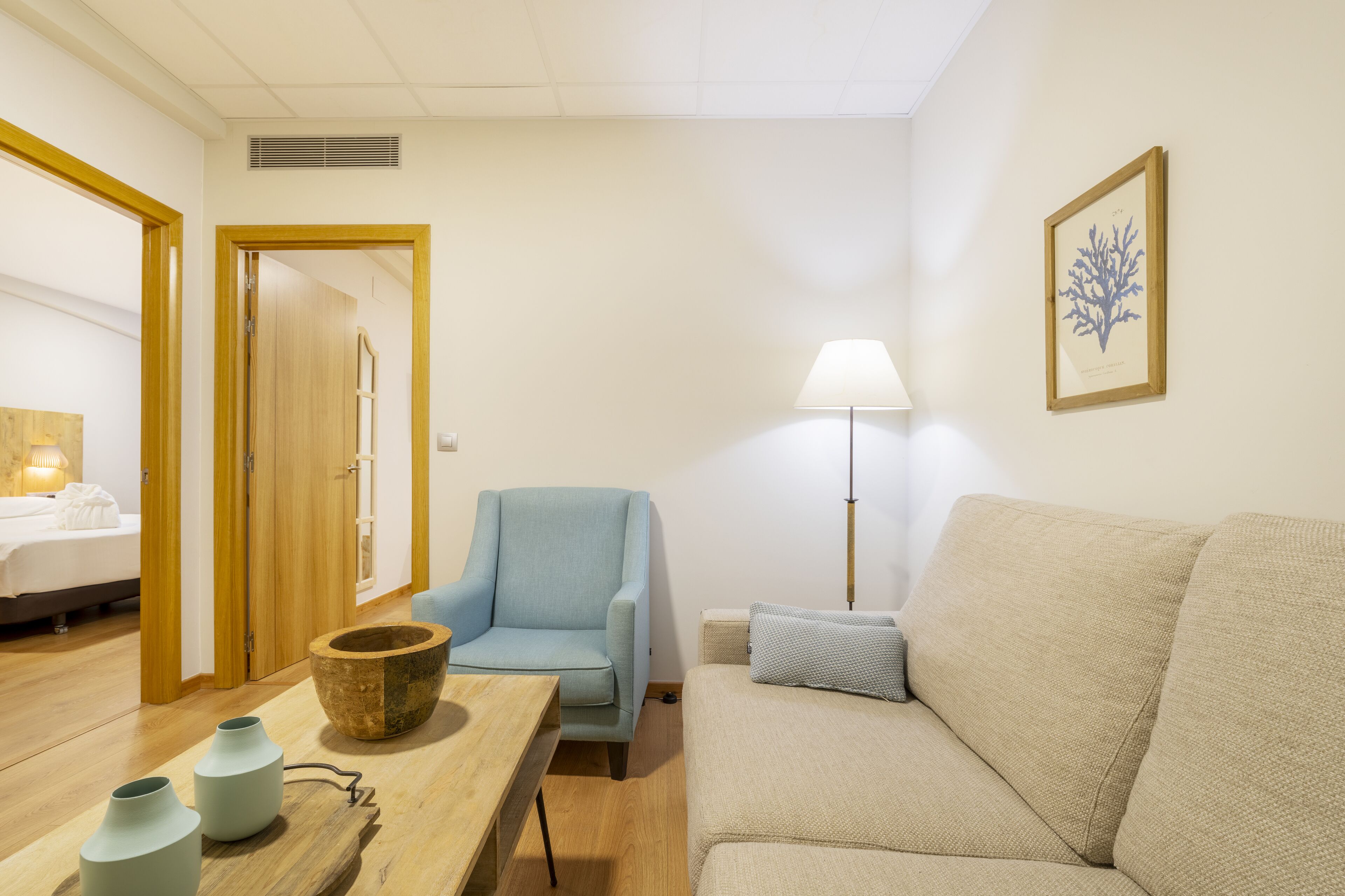 A room with a bed, a blue armchair, a beige sofa, a wooden table with vases, and a floor lamp.