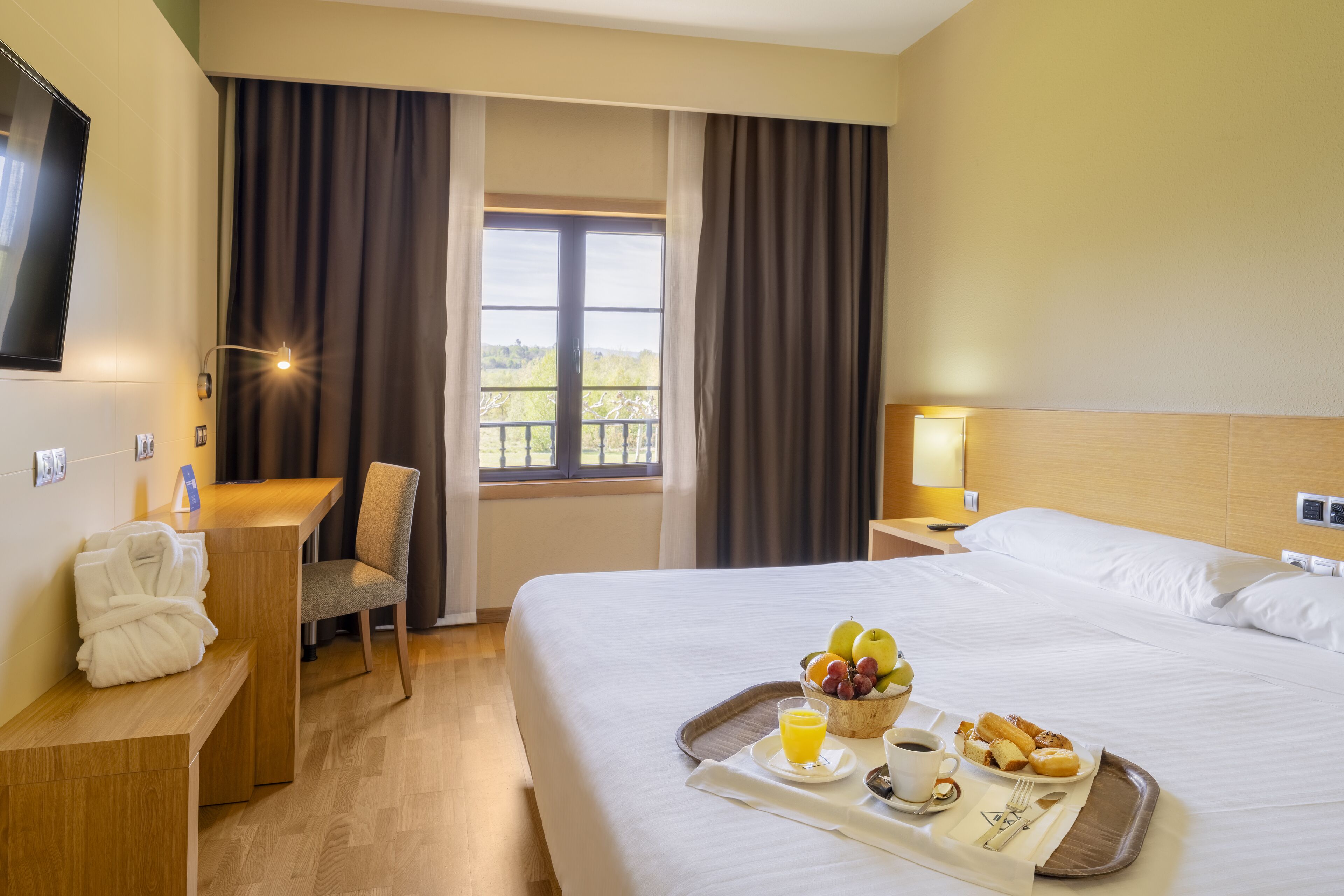 A hotel room with a bed, a desk, a chair, a TV, a window with curtains, and a tray with breakfast items.