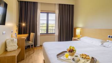 A hotel room with a bed, a desk, a chair, a TV, a window with curtains, and a tray with breakfast items.