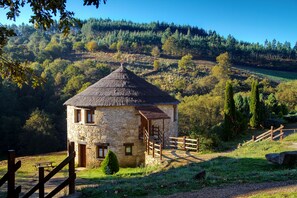 Una cabaña de piedra con techo de paja, un porche de madera y vista a una ladera boscosa.