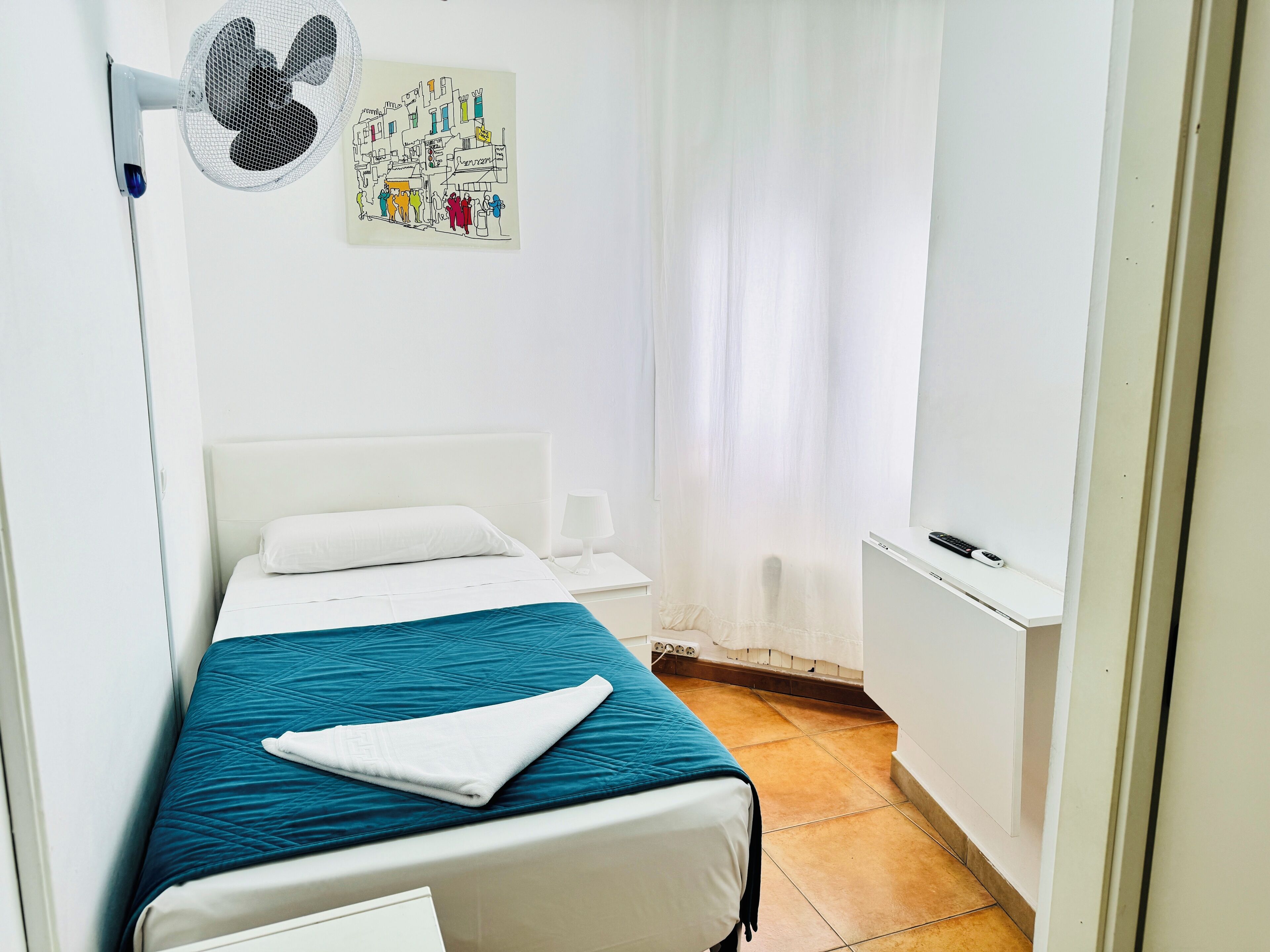 A bedroom with a bed, a bedside table, a lamp, a wall-mounted fan, and a window with curtains.