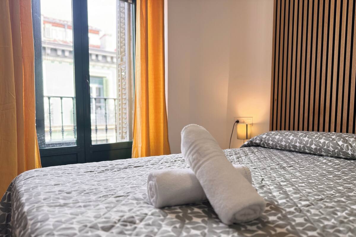 A hotel room with a bed, a window with curtains, a bedside lamp, and a view of buildings outside.