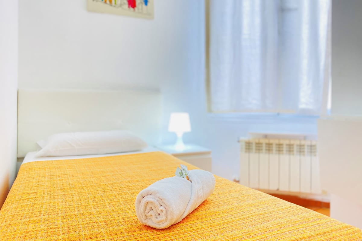 A neatly made bed with a rolled white towel, a white pillow, and a yellow textured bedspread in a bright room with a white headboard, a window with sheer curtains, and a radiator.