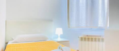 Cama bem arrumada com uma toalha branca enrolada, um travesseiro branco e um edredom amarelo texturizado num quarto claro com cabeceira branca, janela com cortinas leves e radiador.