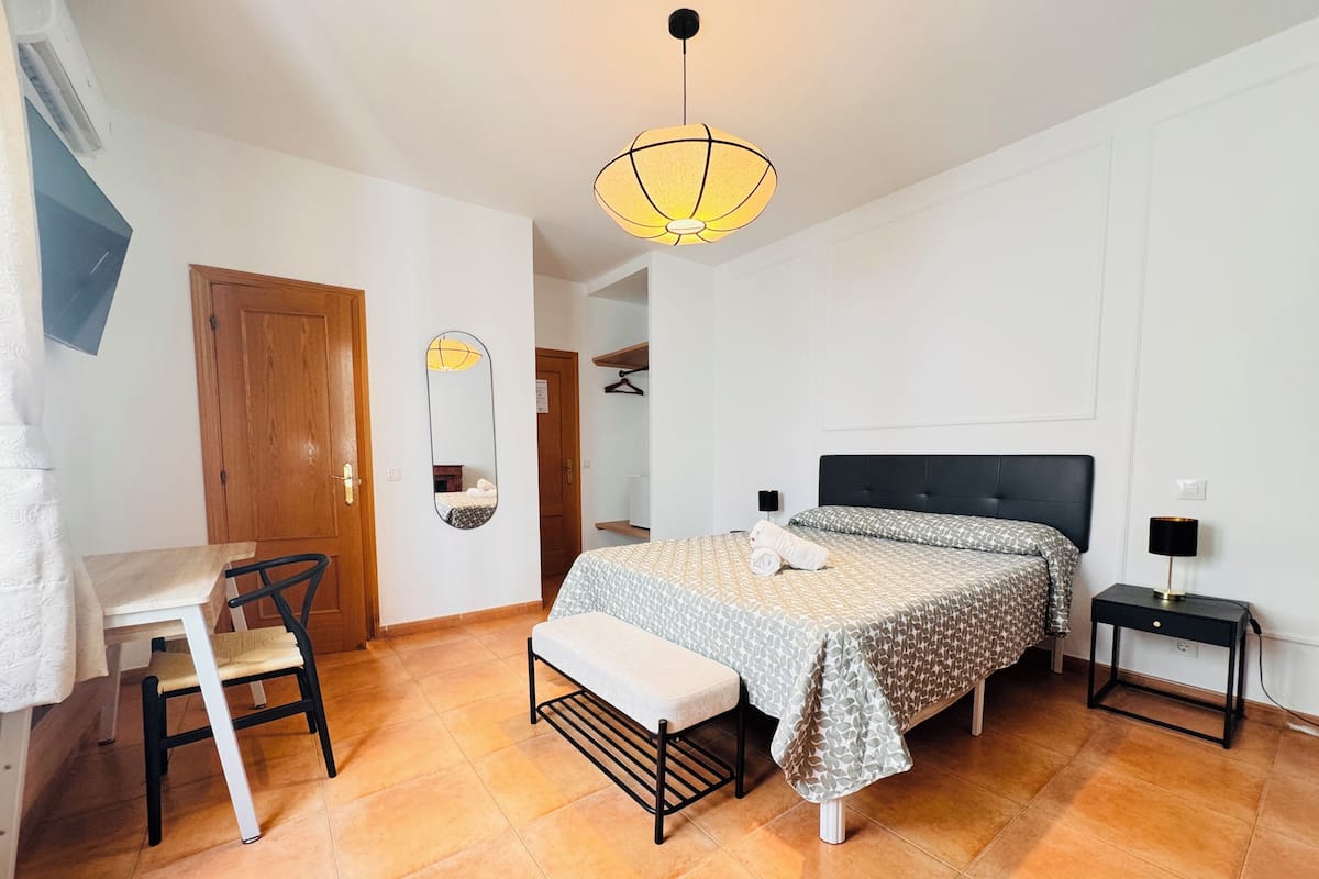 A bedroom with a bed, a desk, a chair, a bedside table, and a hanging light fixture.