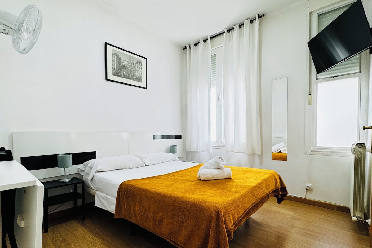 A hotel room with a bed, a desk, a chair, a window with curtains, a wall-mounted TV, and a framed picture.