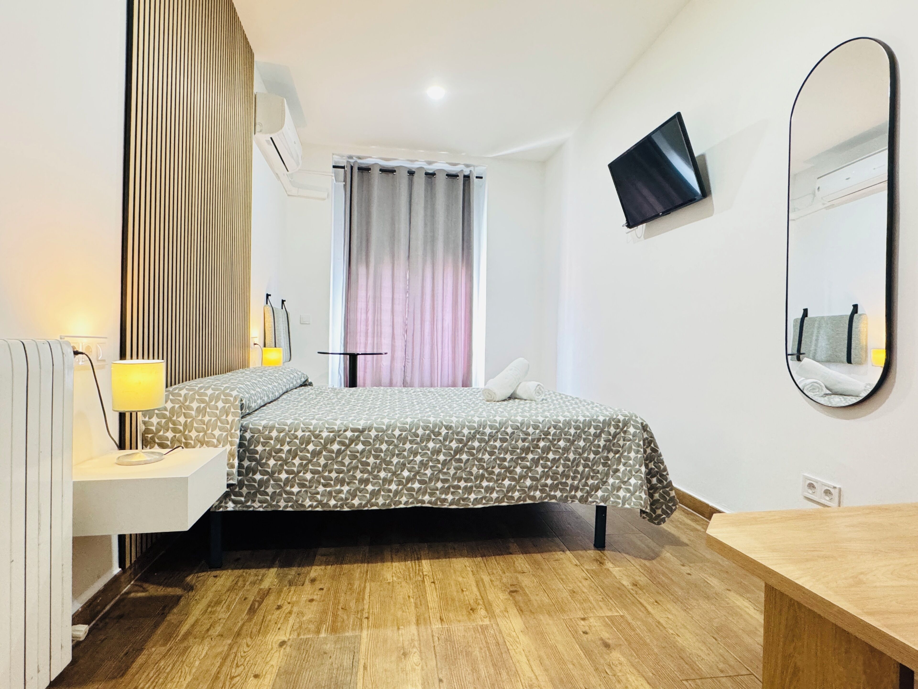 A hotel room with a bed, bedside table, lamp, air conditioning unit, window with curtains, and a mounted TV.