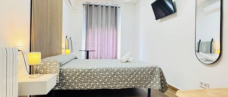 A hotel room with a bed, bedside table, lamp, air conditioning unit, window with curtains, and a mounted TV.