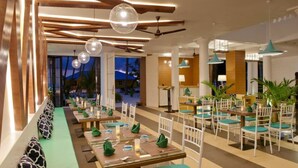 A modern restaurant with long communal tables, pendant lighting, and a view of the outdoors. - Crystal Sands (Maafushi)