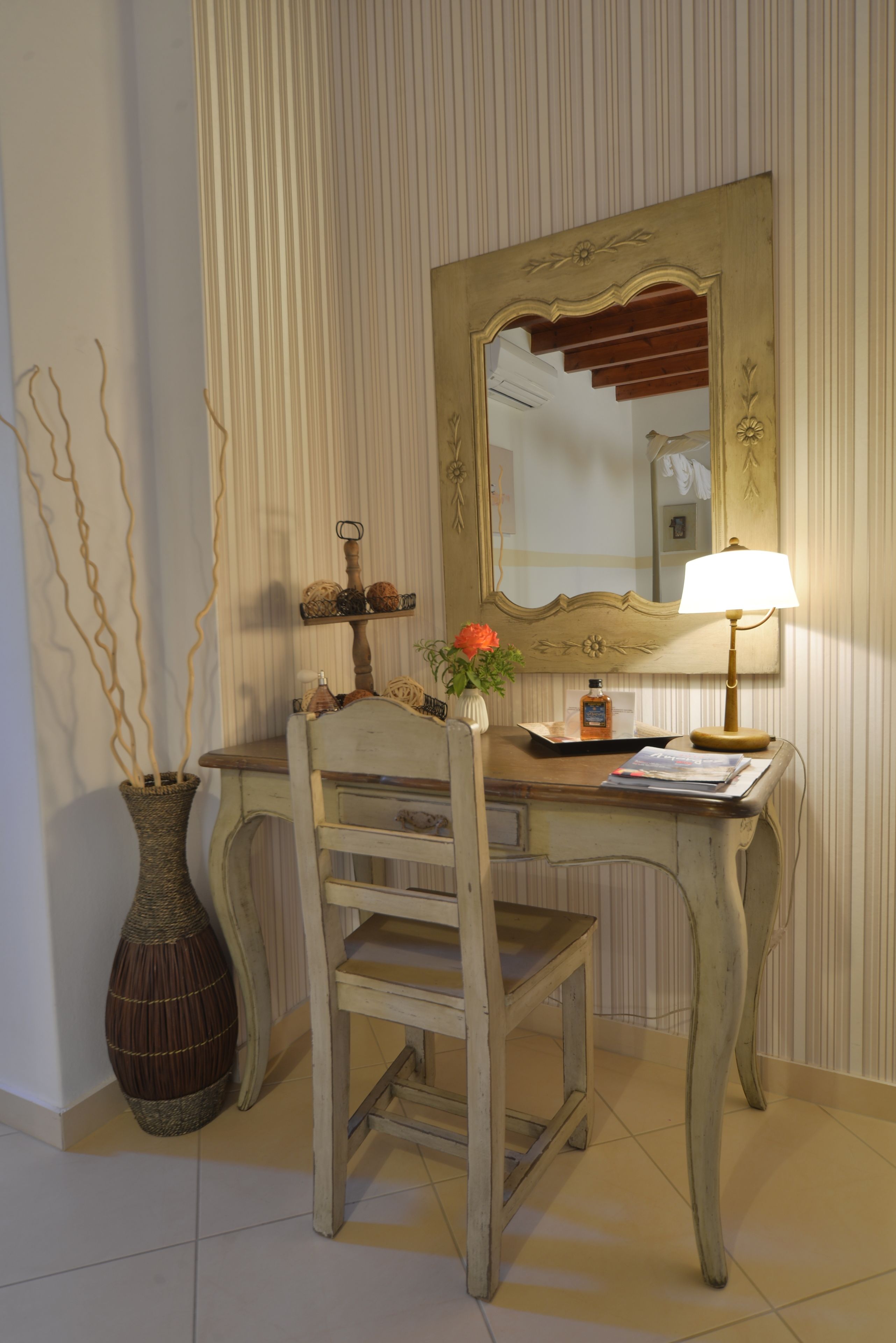 A wooden desk with a mirror, a vase with flowers, a lamp, and a chair.