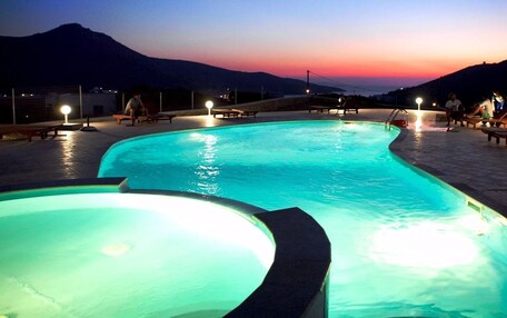 A swimming pool with a curved edge, surrounded by lounge chairs and tables, set against a mountainous backdrop during twilight.