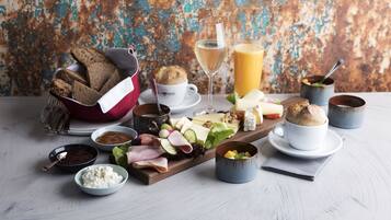 Ein Frühstücksarrangement mit Brot, Käse, Schinken und verschiedenen Aufstrichen auf einer Holzplatte, dazu Getränke und kleine Schälchen mit Gewürzen.