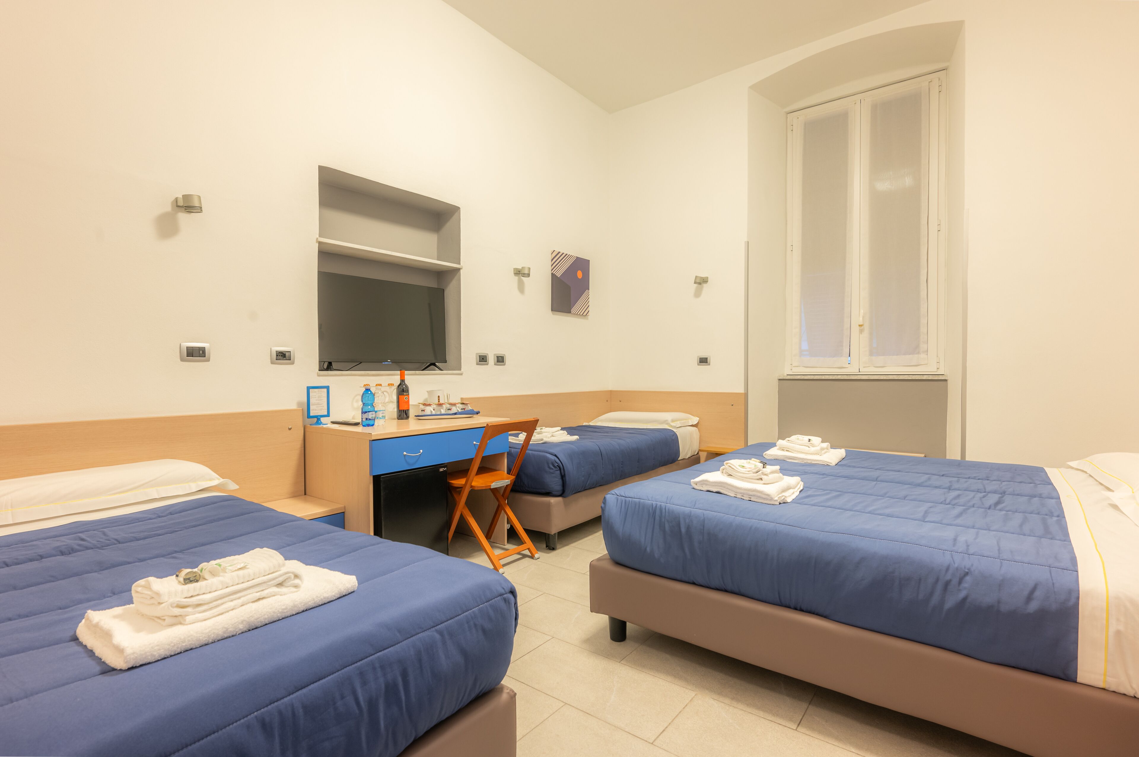 A hotel room with two beds, a desk, a chair, a TV, and a window.