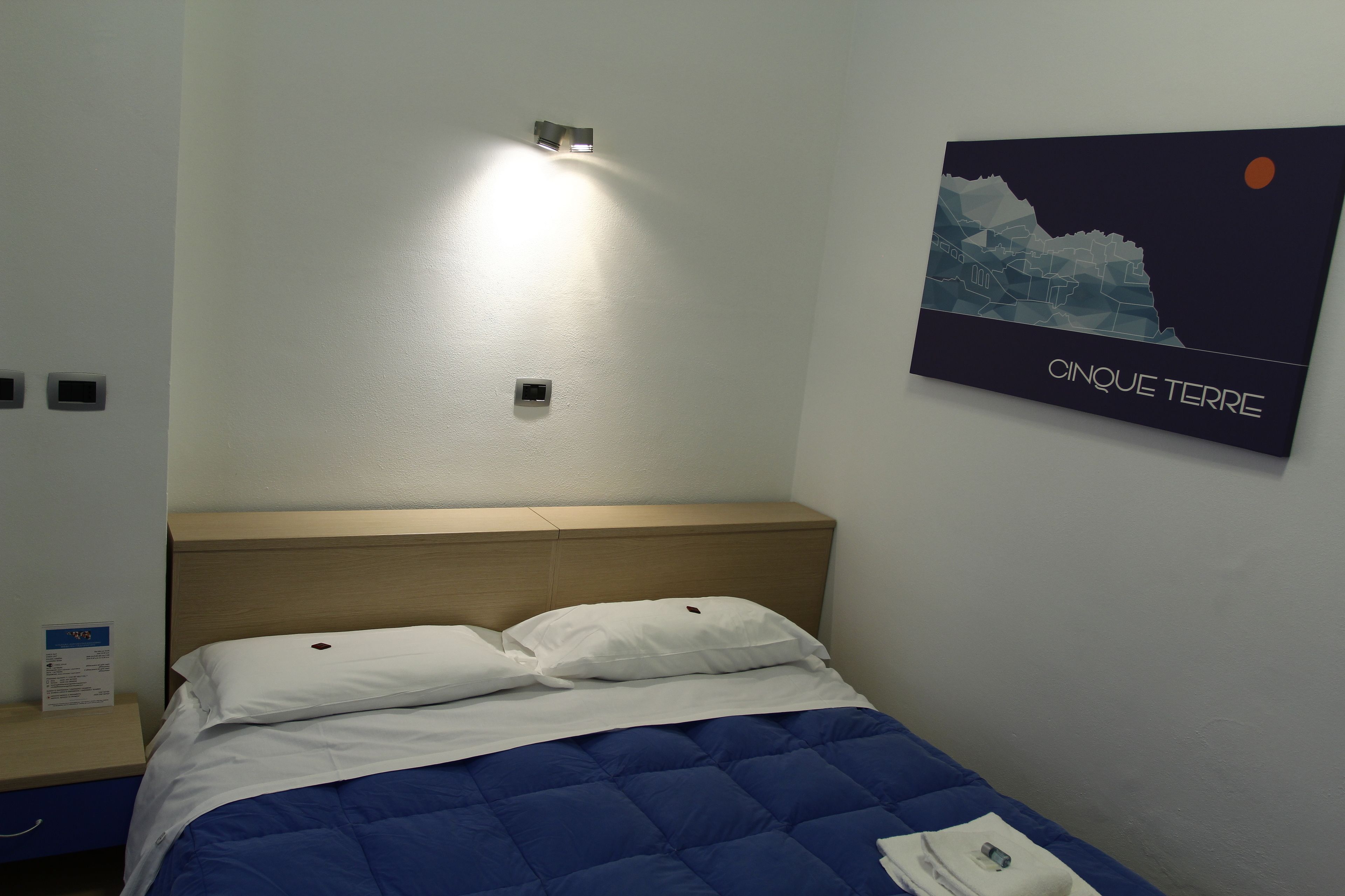 A hotel room with a bed, a headboard, a wall-mounted light, and a poster of a cityscape.