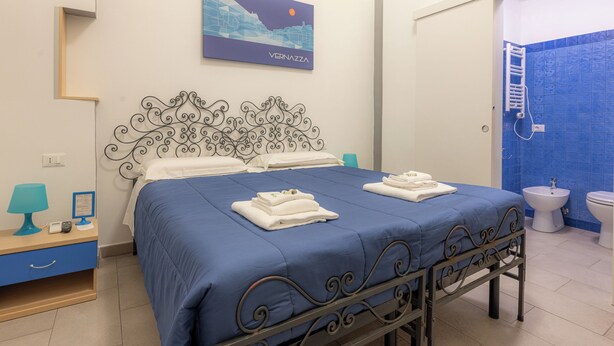 A hotel room with a bed, a desk with a lamp, a bathroom with a blue-tiled wall, and a painting on the wall. - Affittacamere del Golfo e delle Cinque Terre (La Spezia)