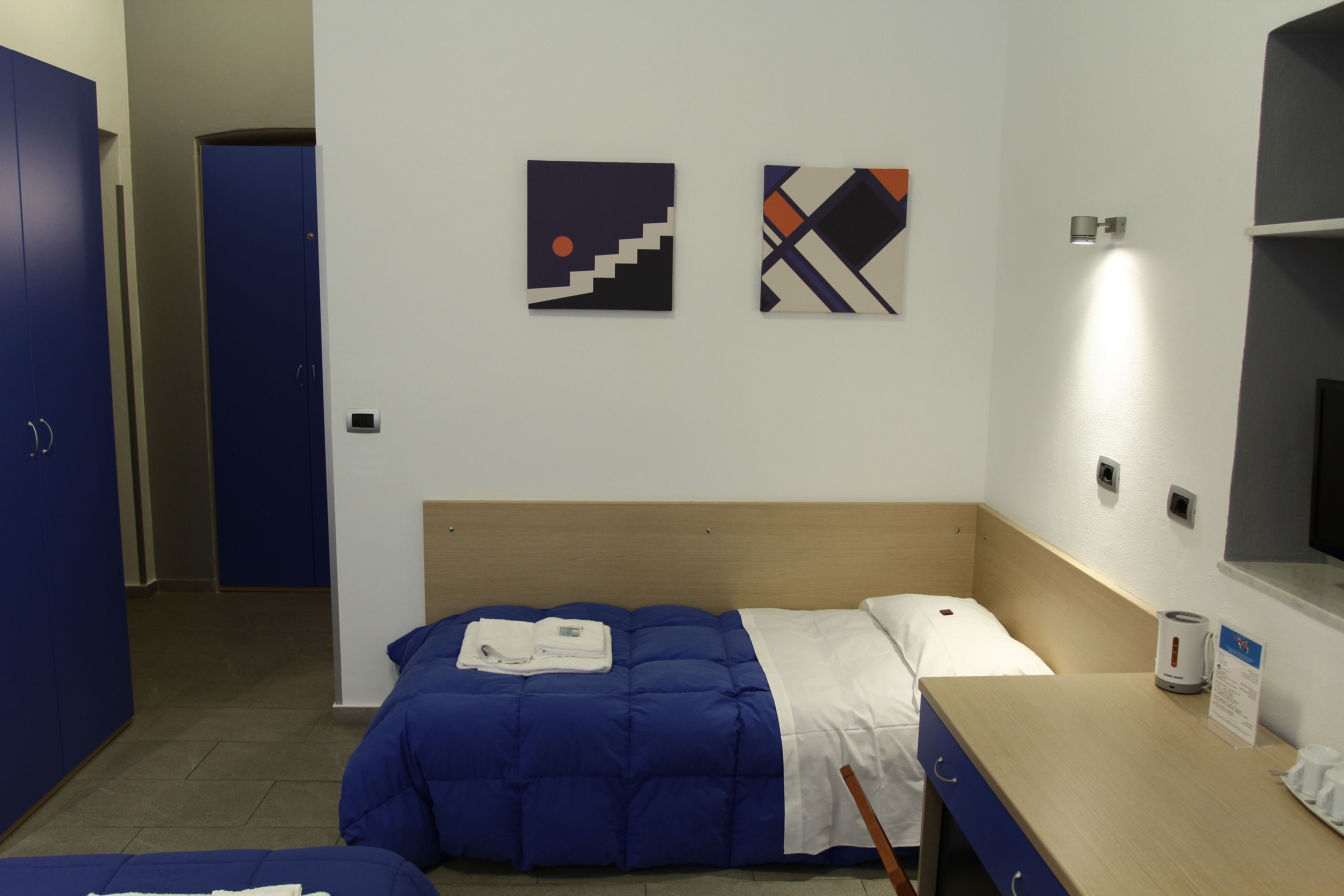 Habitación de hotel con cama, escritorio y silla. Hay dos cuadros en la pared y una puerta al fondo.