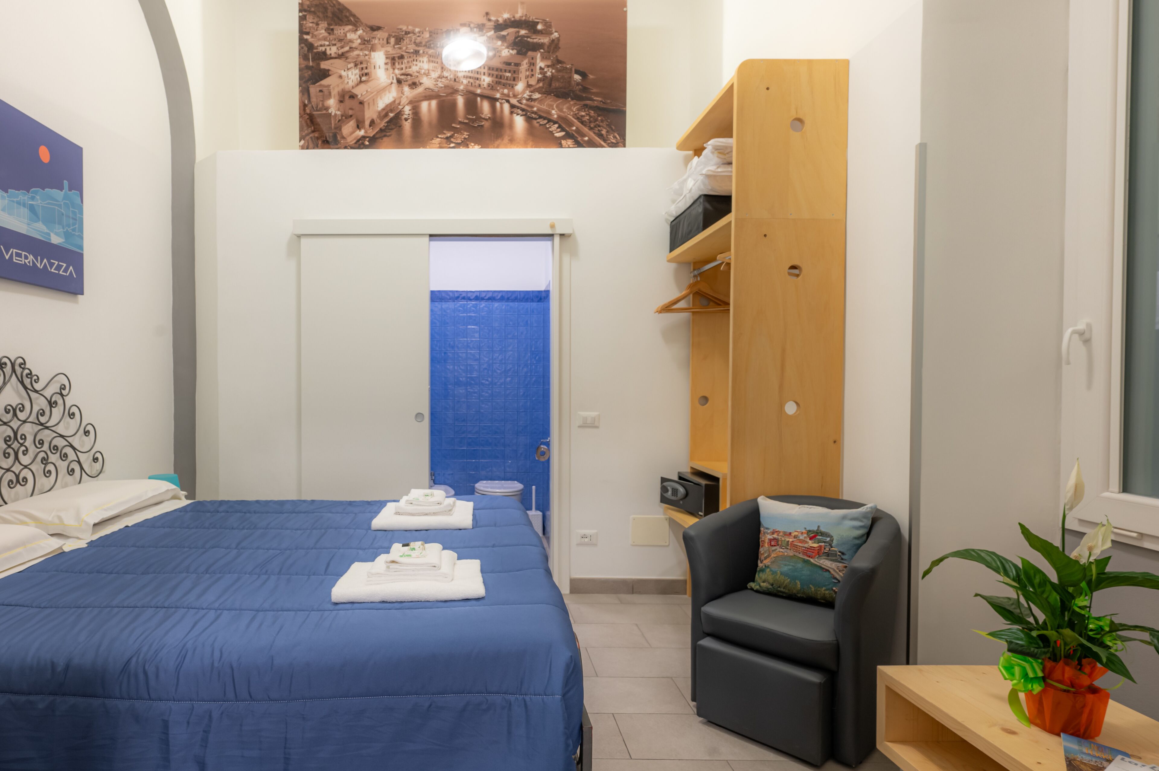Une chambre d’hôtel avec un lit, une chaise, une petite table, une plante et un mur orné d’un tableau représentant un paysage urbain.