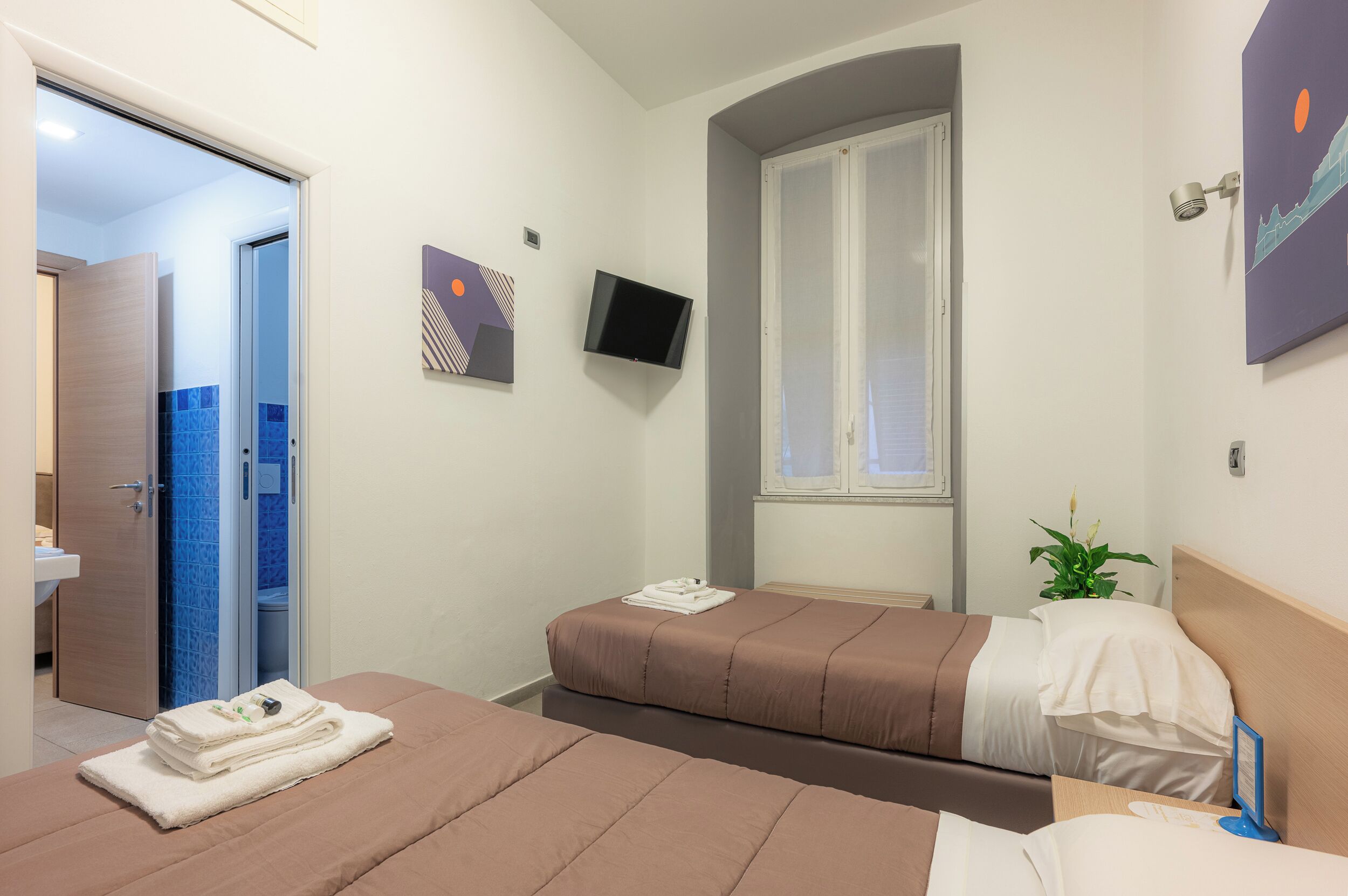 Habitación de hotel con dos camas, televisión, ventana y baño visible a través de una puerta abierta.