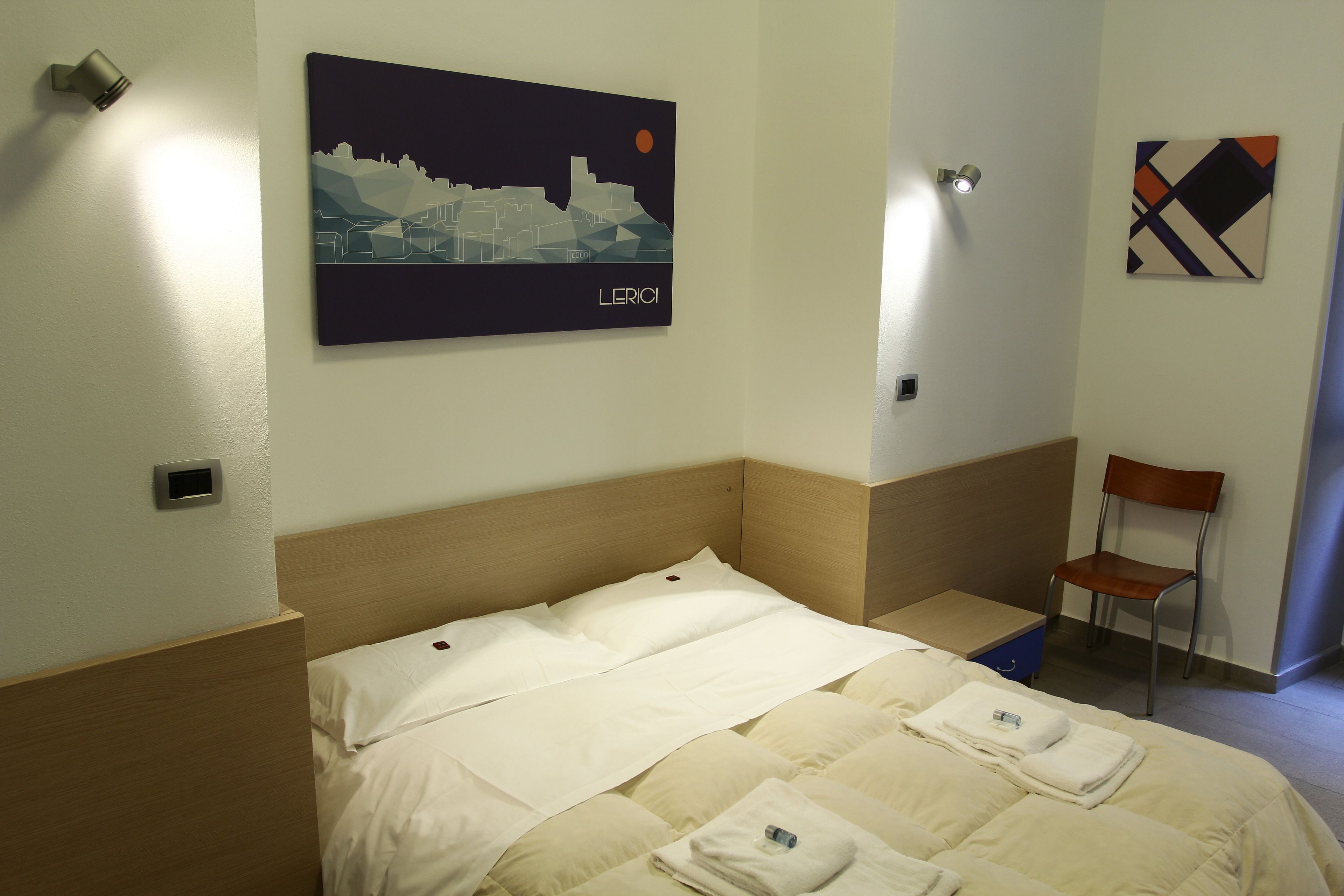 Quarto de hotel com cama, uma cadeira, uma luminária na parede e uma pintura de paisagem urbana.