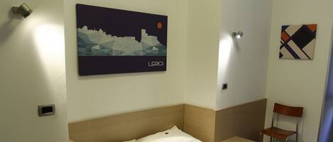 Quarto de hotel com cama, uma cadeira, uma luminária na parede e uma pintura de paisagem urbana.
