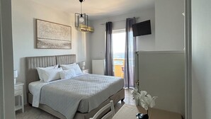 A hotel room with a bed, a bedside table, a flat-screen TV mounted on the wall, and a view of the outdoors through a window. - Eliza Apartments (Rethymno)