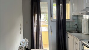 A kitchen with white cabinets, a sink, and a window with blinds. There is a table with a vase of flowers and a hanging light fixture. - Eliza Apartments (Rethymno)