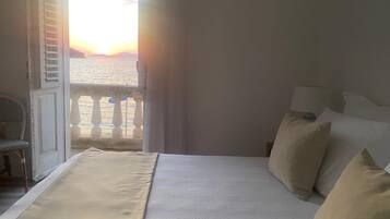 Ein Schlafzimmer mit Bett, Kissen, einem Nachttisch, einem Sessel und Blick auf das Meer bei Sonnenuntergang.