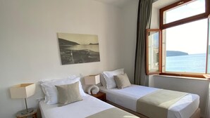 Camera d'albergo con due letti, una finestra vista mare, un quadro appeso al muro e una lampada su un comodino.