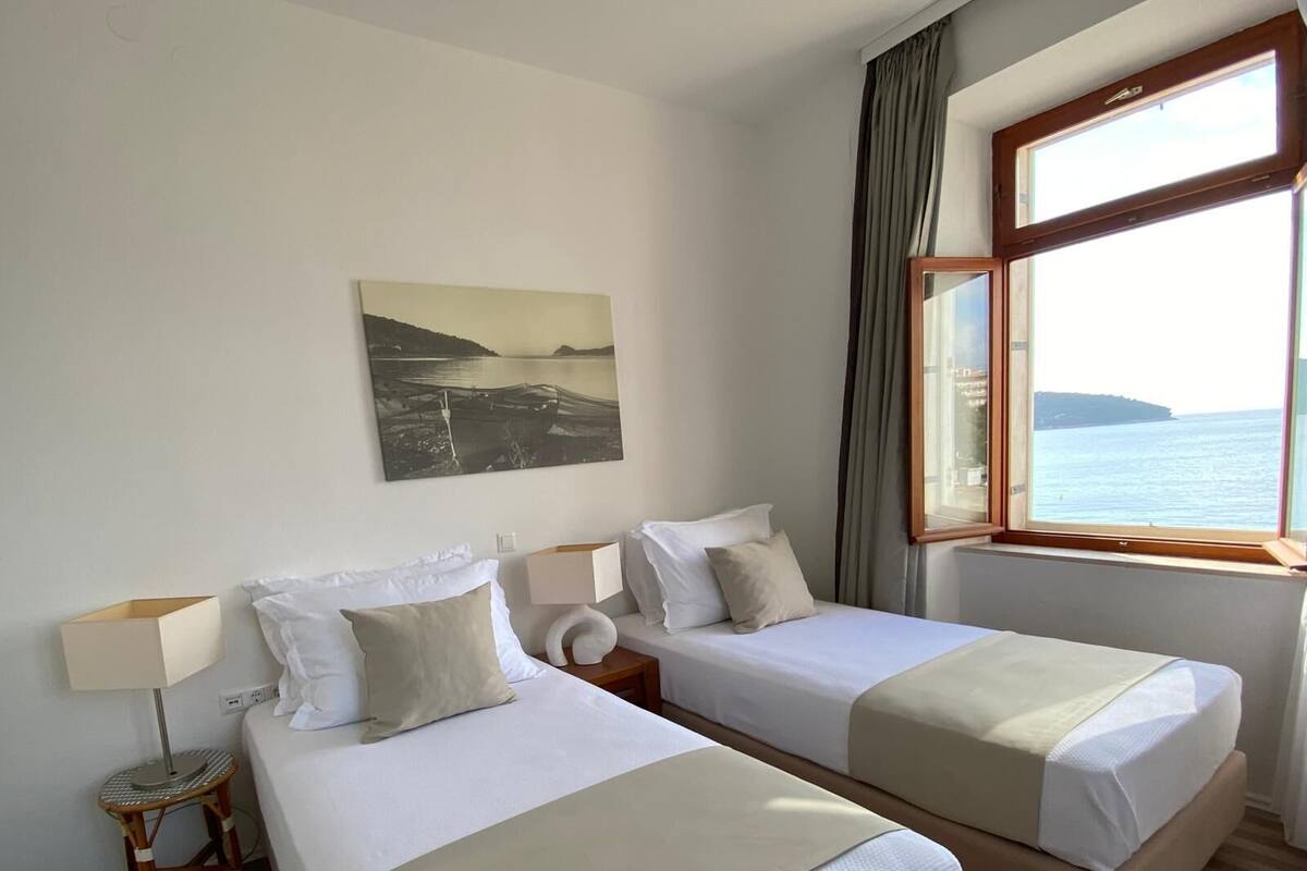 A hotel room with two beds, a window with a view of the sea, a painting on the wall, and a lamp on a side table.
