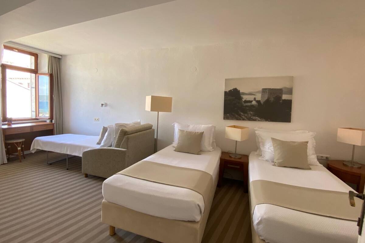 A hotel room with two beds, a desk, and a chair. There is a window with curtains, a painting on the wall, and two bedside lamps.