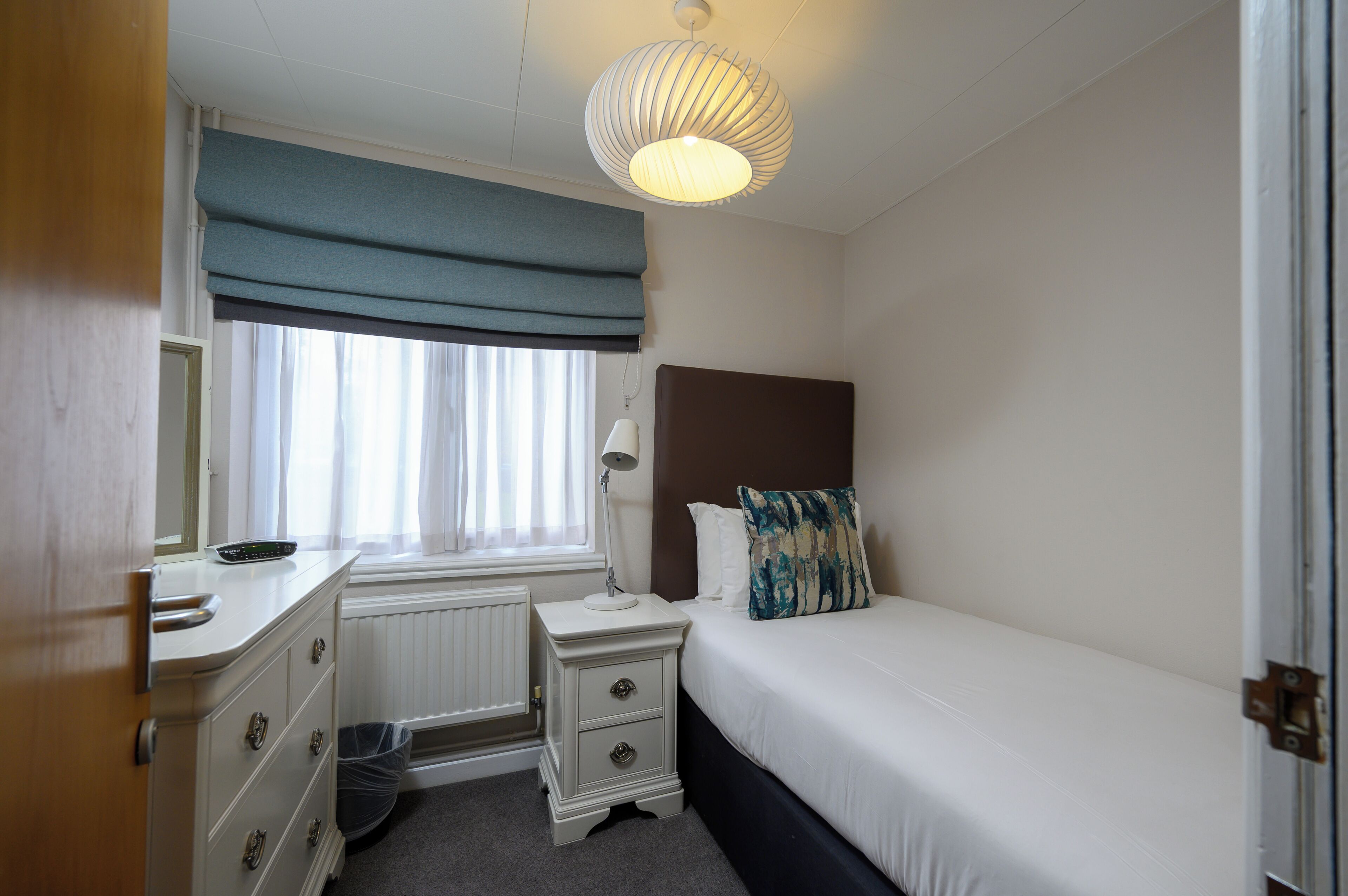 Une chambre à coucher avec un lit, une commode, une table de chevet, une lampe et une fenêtre avec des rideaux.
