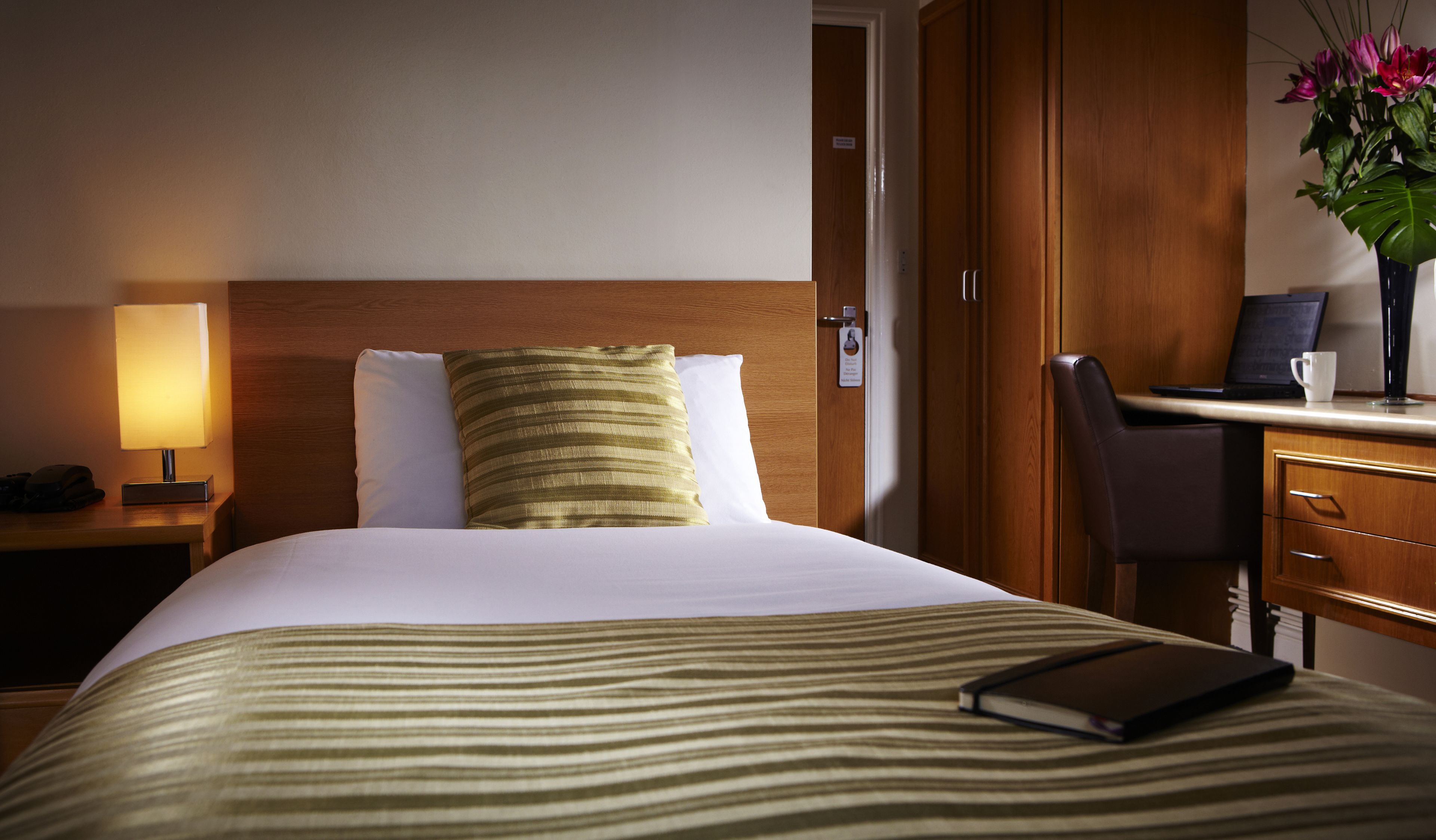 Quarto de hotel com uma cama, uma secretária com um laptop, um telefone e um vaso com flores.