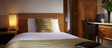 Quarto de hotel com uma cama, uma secretária com um laptop, um telefone e um vaso com flores.