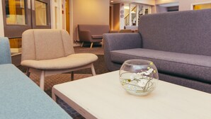 A modern lounge area with seating arrangements including a grey sofa and a beige chair. A clear glass bowl with white flowers is placed on a white table. - Peter Scott House, Birmingham (Birmingham)