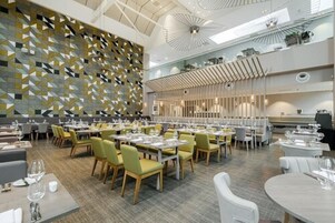 A spacious dining area with multiple tables and chairs, a patterned wall, and a high ceiling with modern lighting fixtures.