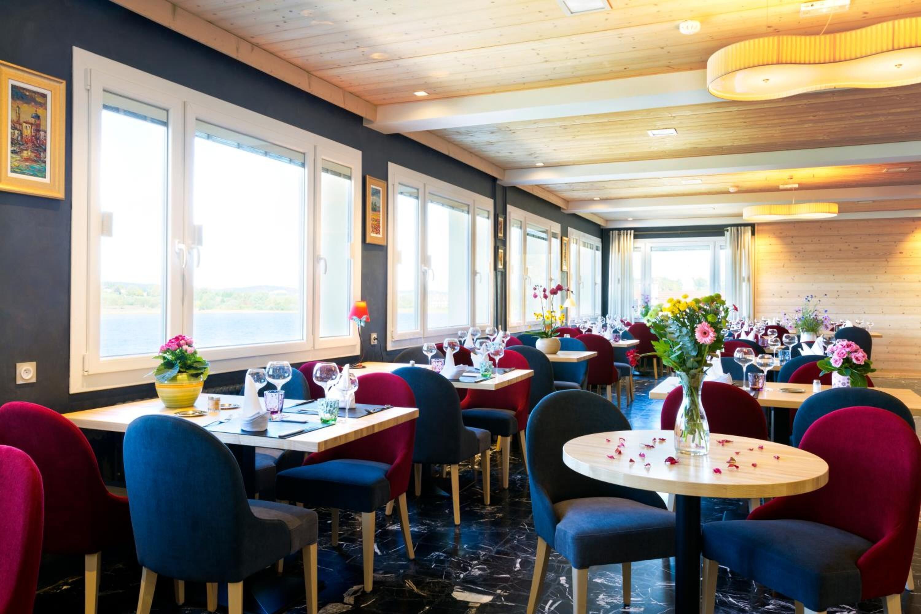 Un restaurant doté de nombreuses tables et chaises, de grandes fenêtres et d’un plafond en bois.
