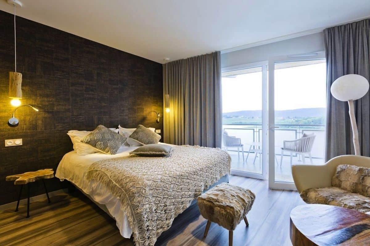 A modern bedroom with a large bed, a balcony with a view, and a seating area with a chair and ottoman.