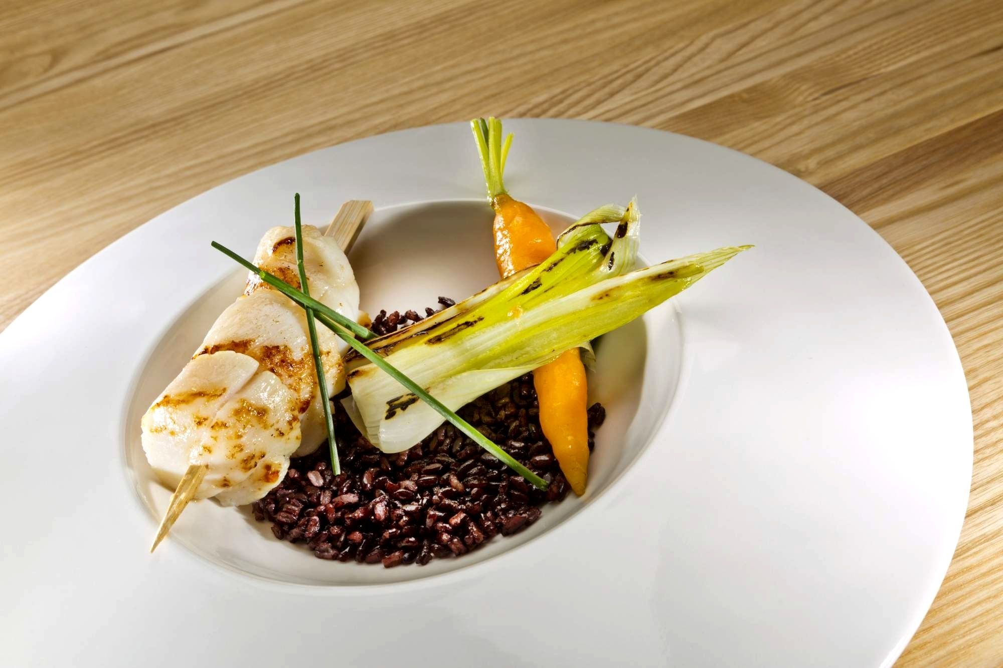 A white plate with a dish containing black rice, a piece of grilled chicken, and a carrot.