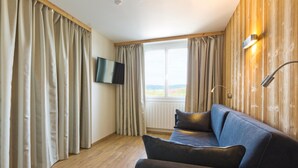 A room with a wooden wall, a large window with curtains, a sofa, a flat-screen TV, and a wall-mounted lamp. - Hotel de l'Abbaye (Grande-Rivière-Château)