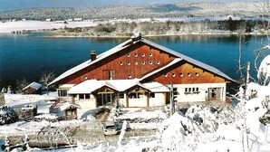 A large wooden building with multiple windows, a covered porch, and a snow-covered landscape with a lake in the background. - Hotel de l'Abbaye (Grande-Rivière-Château)