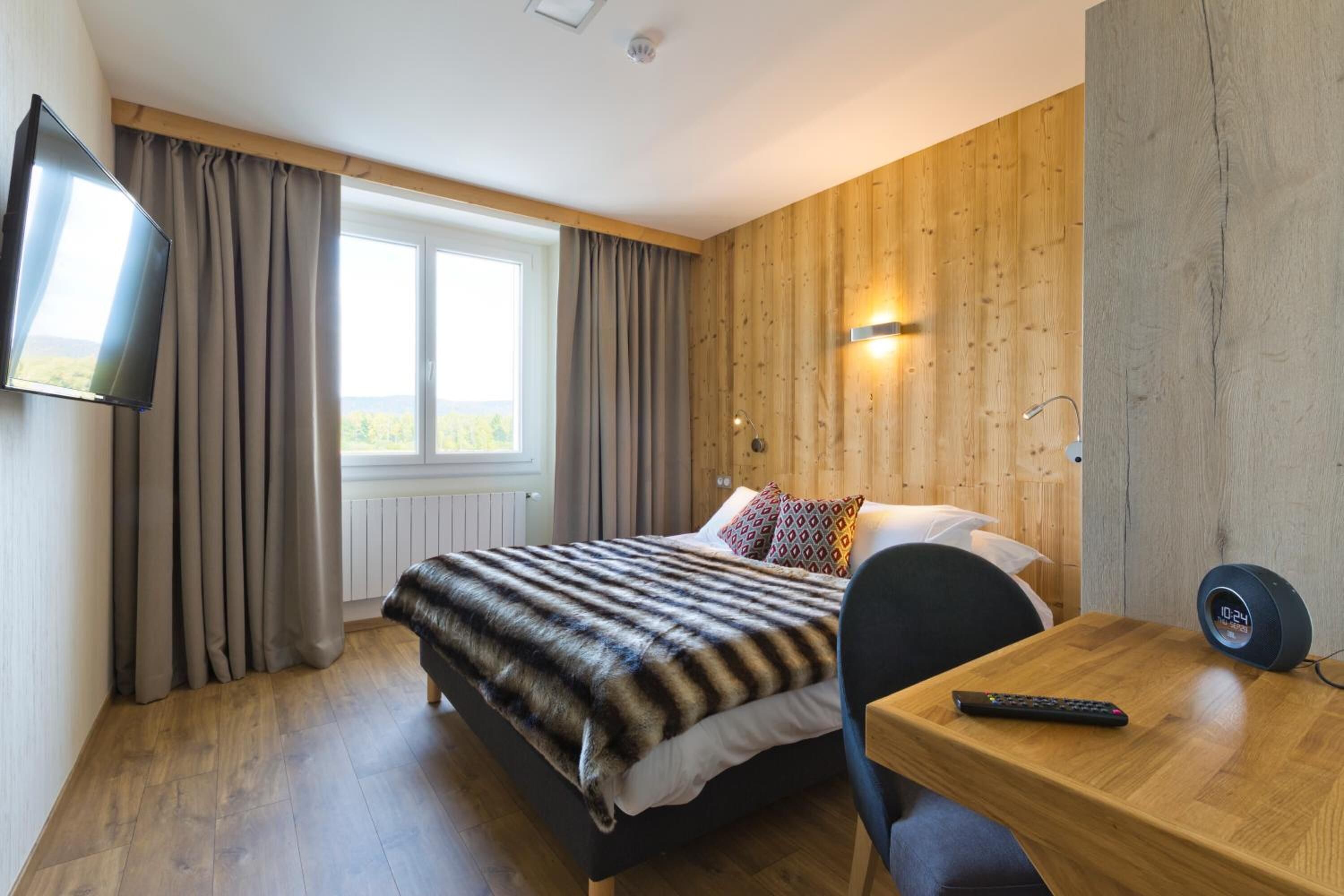A hotel room with a bed, a TV mounted on the wall, a wooden desk, and a window with curtains.