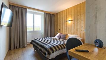 A hotel room with a bed, a TV mounted on the wall, a wooden desk, and a window with curtains.