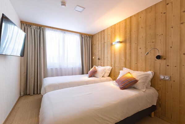 A hotel room with two beds, wooden panel walls, a TV, and a window with curtains.
