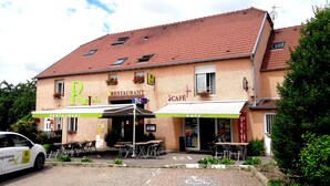 Een gebouw met twee verdiepingen, met een restaurant en café op de begane grond. Er staan terrastafels en -stoelen, en er is een auto geparkeerd voor de deur.