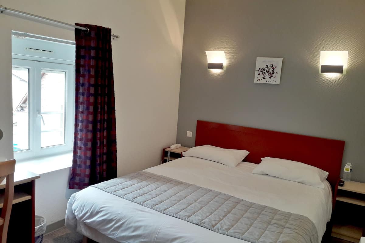 Una cama bien tendida con cabecera roja, una mesita de noche con lámpara y una ventana con cortinas.
