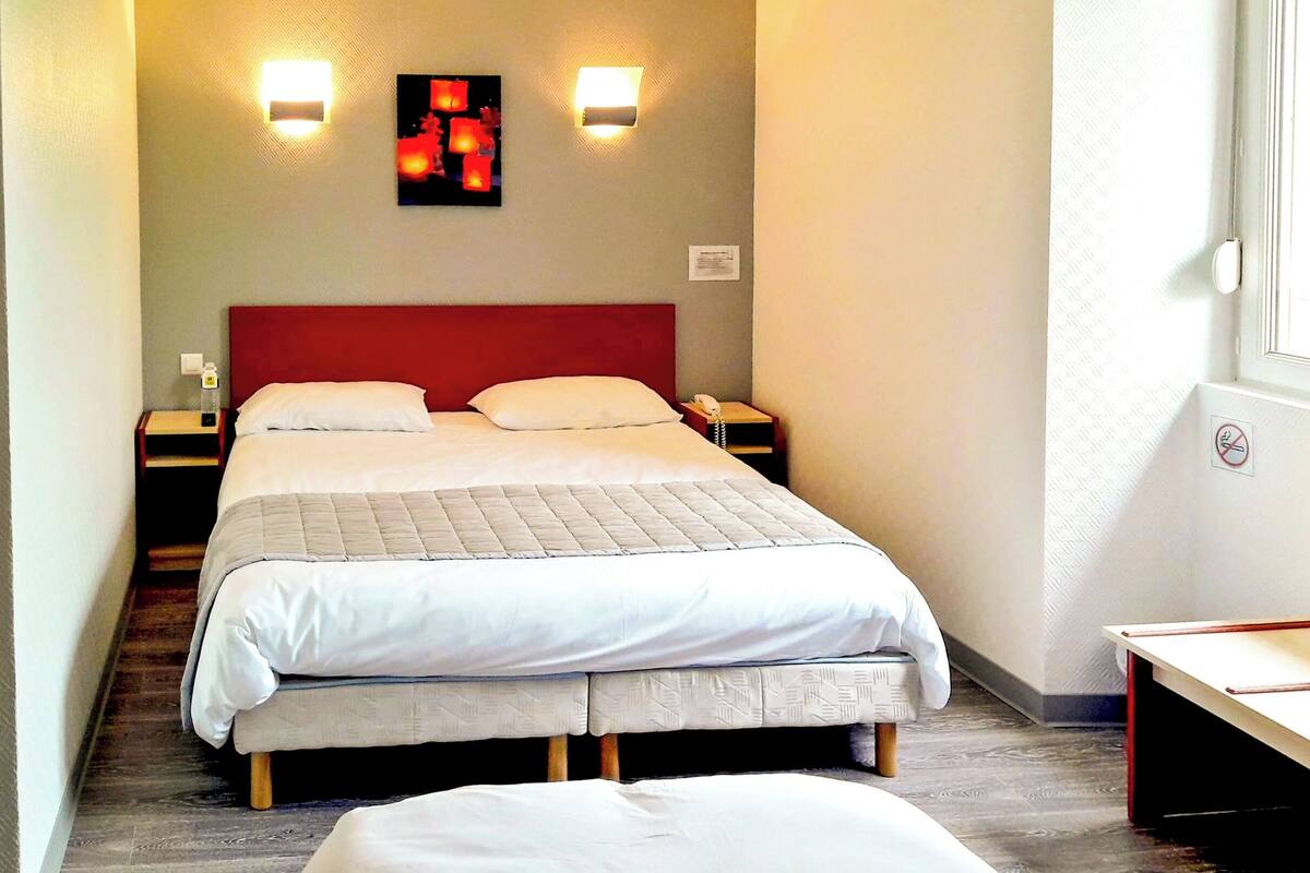 Una habitación de hotel con cama, dos mesitas de noche, lámpara de pared y ventana con vistas.