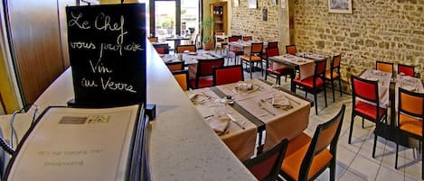 A restaurant with stone walls, tables set with red chairs, and a chalkboard menu.